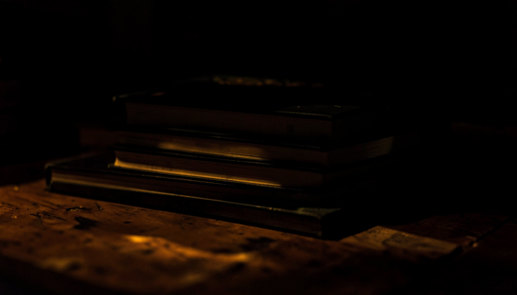 A dark, low-key photograph of stacked hardcover books on a wooden table, deep shadows pooling around them, with a single warm light catching the spines.