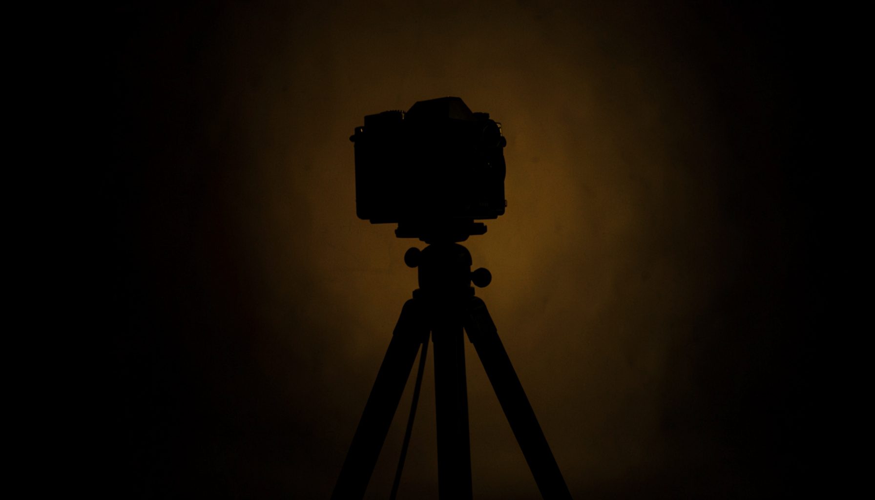 A dark, low-key photograph of a vintage film camera on a tripod, silhouetted against a dimly lit studio backdrop with deep shadows and warm amber highlights.