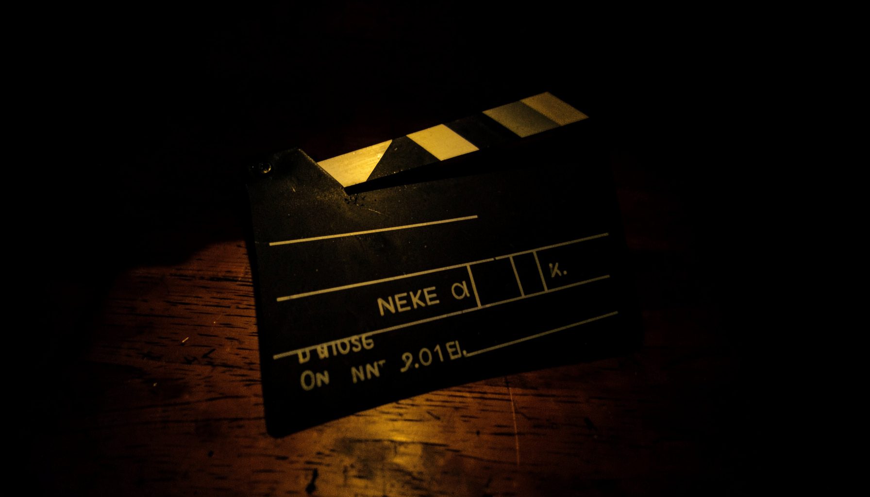 A dark, low-key photograph of a clapperboard resting on a wooden surface, lit by a single warm practical light, with deep shadows pooling around it.
