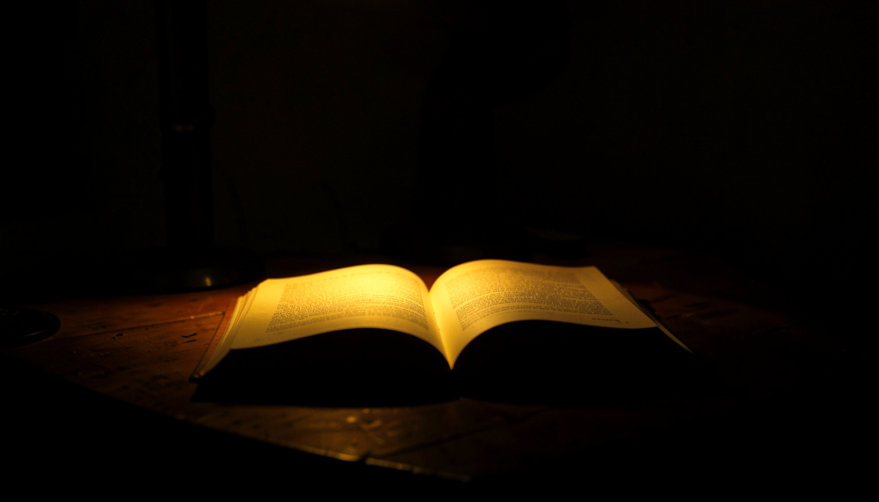 Dark photograph of an open book resting on a wooden desk, lit by a single warm lamp in a shadow-filled room.