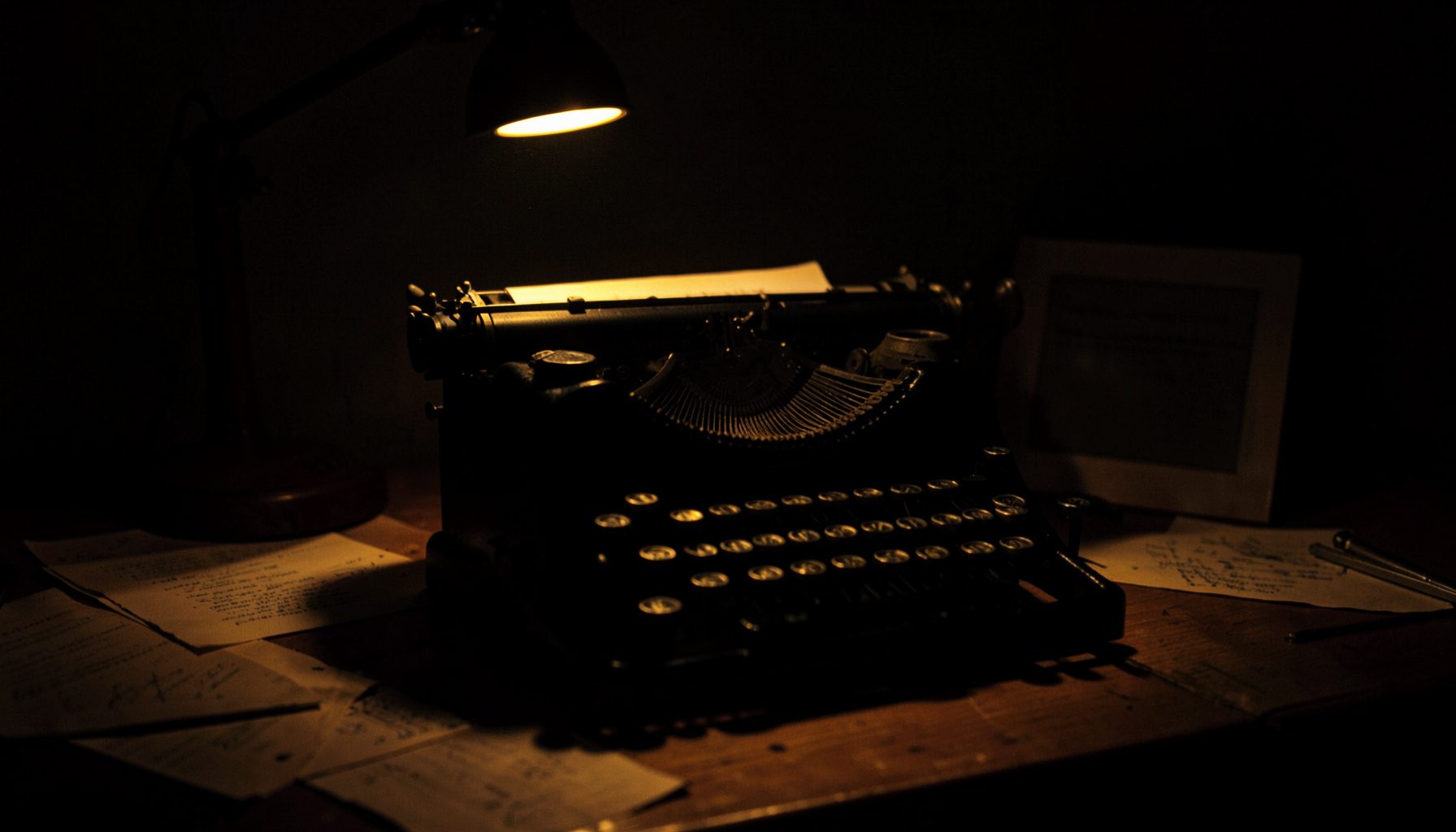 A dark, low-key photograph of a vintage typewriter on a wooden desk, lit by a single warm desk lamp casting deep shadows across scattered pages.